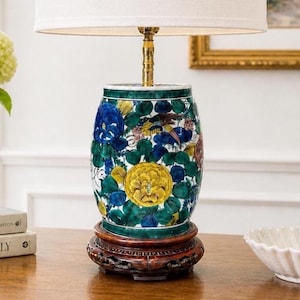 May include: A decorative table lamp with a white lampshade and a ceramic base. The base features a floral design with blue, yellow, and green accents. The lamp sits on a dark wooden base, adding a touch of elegance to any room.