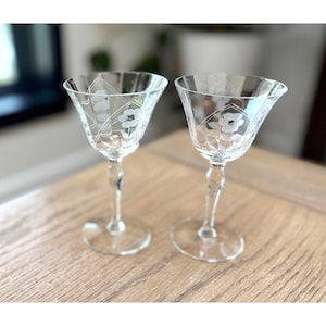 May include: Two clear, stemmed wine glasses with etched floral designs. The glasses have a flared bowl shape and sit on a round base. The etched design includes flowers and delicate lines. The glasses are displayed on a light brown wooden surface.