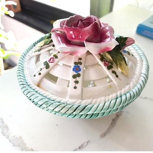 May include: A round ceramic lidded bowl featuring a white lattice-patterned lid topped with a large, sculpted pink rose and green leaves. The bowl has a light green rope-textured rim and is decorated with small hand-painted floral motifs.