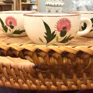 May include: Three cream-colored teacups with a pink thistle design and green leaves sit on a round, woven wicker tray. The cups have brown rims and handles. The tray is made of woven, light brown wicker.