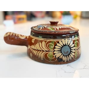 May include: A small, round, brown ceramic pot with a lid and handle. The pot is decorated with white, green, and blue floral patterns. The lid has a small knob on top. The pot is likely used for cooking or serving food.