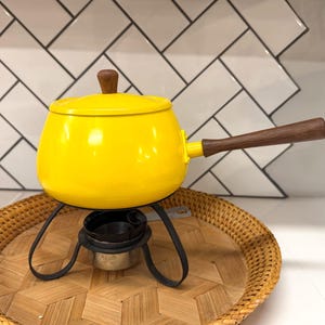 May include: A bright yellow fondue pot with a wooden handle and lid knob. The pot sits on a black metal stand above a small burner, placed on a woven tray. The background features a white tile pattern.