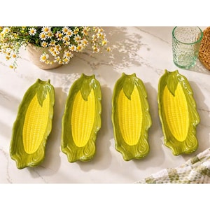 May include: Four ceramic corn-shaped serving dishes. Each dish features a yellow corn cob design with green husks. The dishes are arranged on a marble surface, with a vase of daisies and a glass in the background.