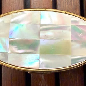 May include: An oval-shaped, vintage trinket box with a gold-colored metal frame. The lid is inlaid with rectangular pieces of iridescent mother-of-pearl, arranged in a checkerboard pattern. The box is set against a wooden surface.