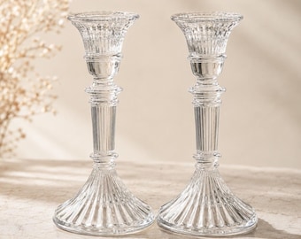 May include: Two clear glass candle holders. Each has a flared base with vertical ridges, a fluted stem, and a decorative top. The candle holders are designed to hold a standard size candle.