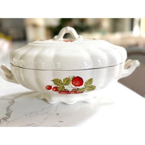 May include: A white ceramic tureen with a scalloped, ribbed body and a matching lid featuring a central handle. The exterior is decorated with a botanical illustration of red strawberries and green leaves on the front and top of the lid.