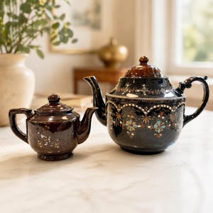 May include: Two decorative teapots. The larger teapot is black with floral accents in blue, gold, and white. The smaller teapot is brown with a matching lid. Both teapots have spouts and handles.