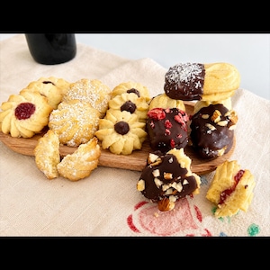May include: An assortment of butter cookies on a wooden board, including some with red jam centers, others dusted with powdered sugar, and several dipped in dark chocolate with toppings like shredded coconut, dried berries, and chopped nuts.