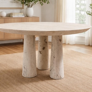 May include: A round, light-colored wooden table with three thick, cylindrical legs. The table sits on a woven rug, and the background includes a wooden cabinet, plants, and a window.