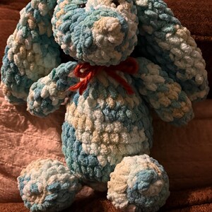 Op de afbeelding: Een handgemaakt pluchen konijn in tinten blauw, wit en beige. Het konijn heeft lange, slappe oren, een rode strik en ronde voetjes. Het zachte, getextureerde garen geeft het konijn een knuffelbaar uiterlijk.