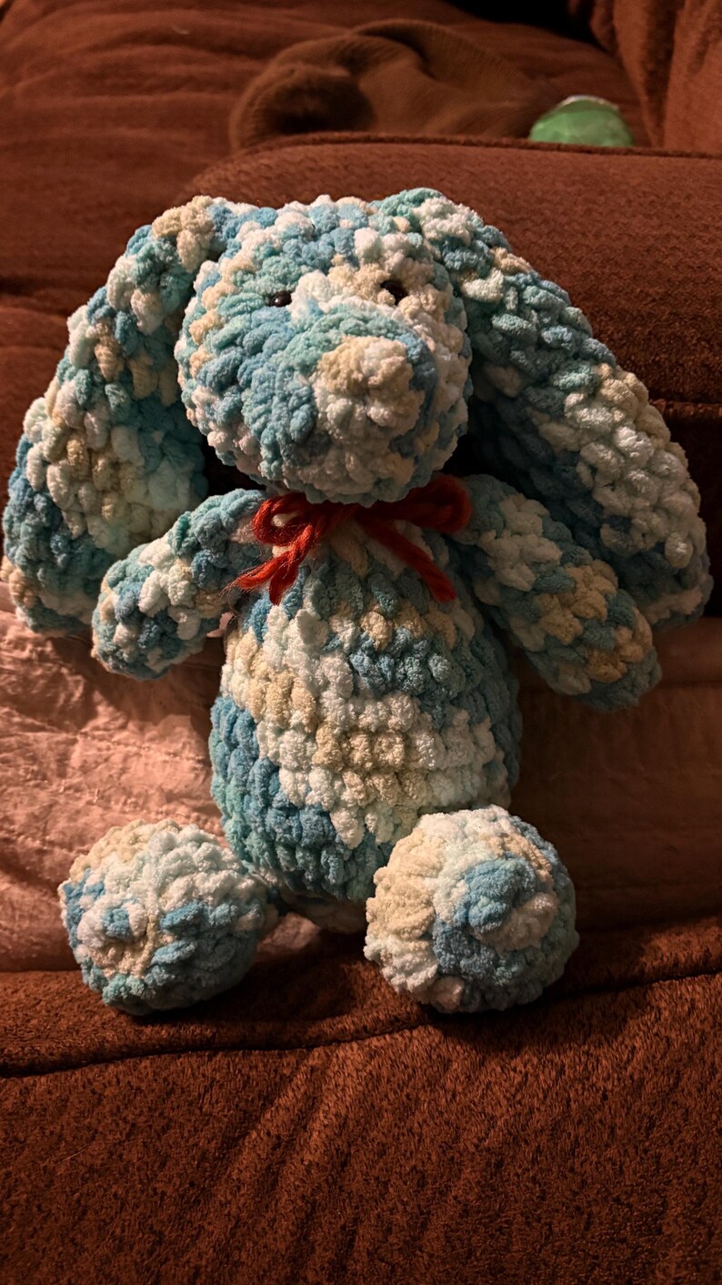 Op de afbeelding: Een handgemaakt pluchen konijn in tinten blauw, wit en beige. Het konijn heeft lange, slappe oren, een rode strik en ronde voetjes. Het zachte, getextureerde garen geeft het konijn een knuffelbaar uiterlijk.