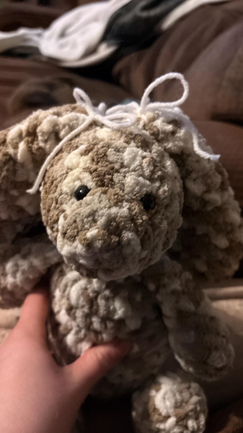 Op de afbeelding: Een handgemaakt, pluche konijn in bruin- en wittinten. Het konijn heeft lange, slappe oren, zwarte ogen en een witte garenstrik. Het zachte, getextureerde materiaal geeft het een knuffelbaar uiterlijk. Het konijn is een knuffel.