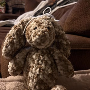 Op de afbeelding: Een handgemaakte gehaakte konijn in bruin- en wittinten. Het pluche speelgoed heeft lange, slappe oren, een kleine witte neus en knoopogen. Het konijn zit op een bruin oppervlak.