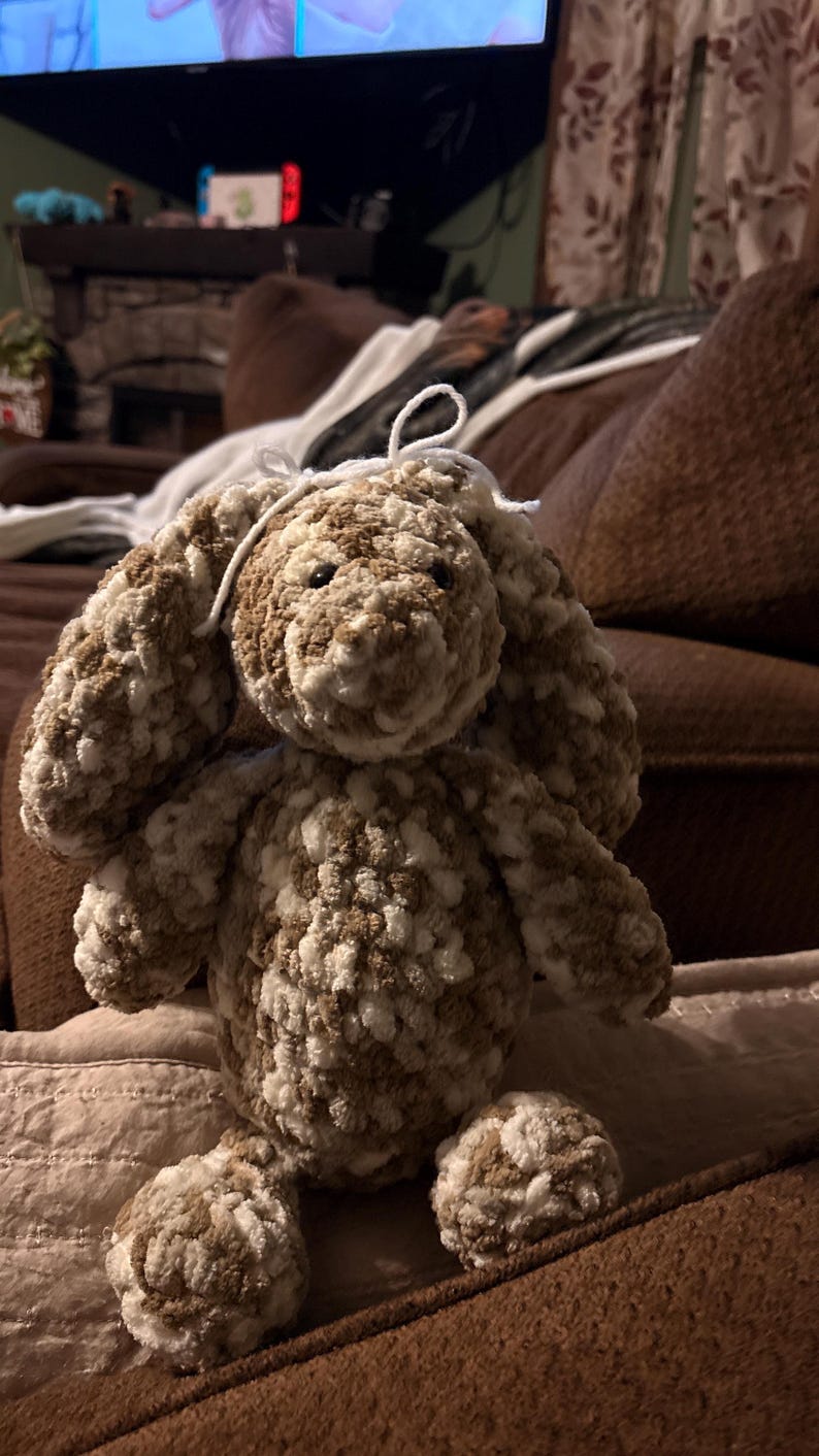 Op de afbeelding: Een handgemaakte gehaakte konijn in bruin- en wittinten. Het pluche speelgoed heeft lange, slappe oren, een kleine witte neus en knoopogen. Het konijn zit op een bruin oppervlak.