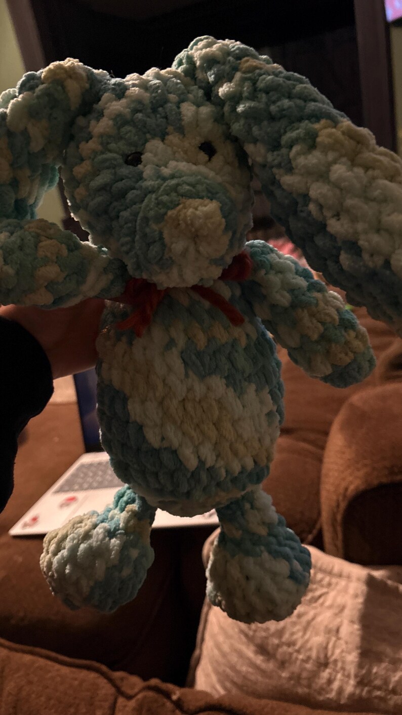 Op de afbeelding: Een handgemaakt pluchen konijn in tinten blauw, wit en geel. Het konijn heeft lange floppy oren, een kleine rode strik en een zacht, getextureerd uiterlijk. Dit knuffelige speelgoed is perfect voor kinderen.