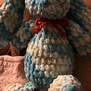 Op de afbeelding: Een handgemaakt pluchen konijn in tinten blauw, wit en beige. Het konijn heeft lange oren, een rond lichaam en een kleine rode strik. Het zachte, getextureerde materiaal geeft het een knuffelbaar uiterlijk. Een charmant, handgemaakt speelgoed.