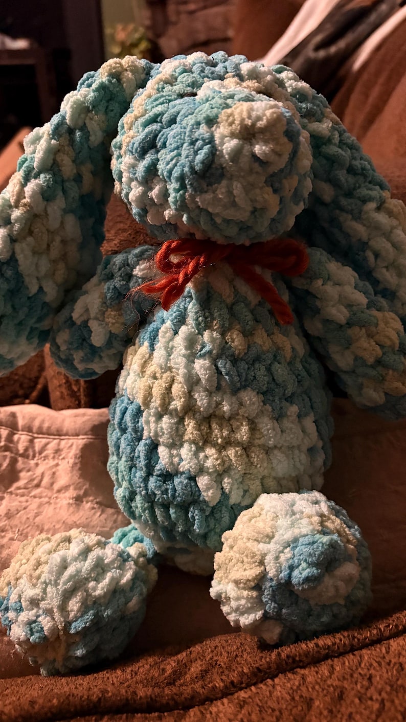 Op de afbeelding: Een handgemaakt pluchen konijn in tinten blauw, wit en beige. Het konijn heeft lange oren, een rond lichaam en een kleine rode strik. Het zachte, getextureerde materiaal geeft het een knuffelbaar uiterlijk. Een charmant, handgemaakt speelgoed.