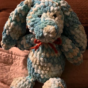 Op de afbeelding: Een handgemaakt konijn van zacht garen in blauwe, witte en beige tinten. Het konijn heeft lange floppy oren, een rode strik om de nek en zwarte ogen. Het zachte, getextureerde garen geeft het een gezellige uitstraling, ideaal als kinderspeelgoed.