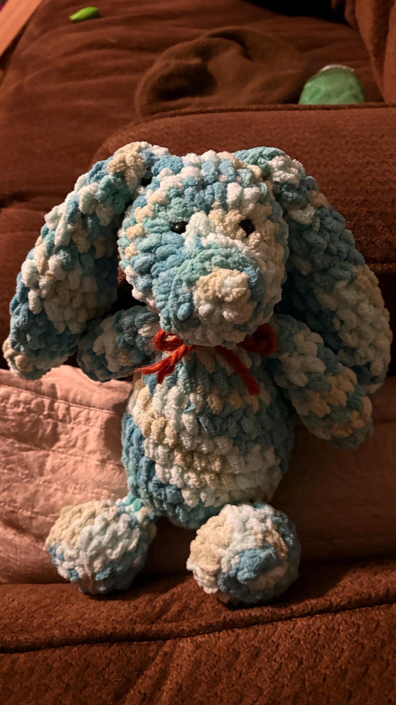 Op de afbeelding: Een handgemaakt konijn van zacht garen in blauwe, witte en beige tinten. Het konijn heeft lange floppy oren, een rode strik om de nek en zwarte ogen. Het zachte, getextureerde garen geeft het een gezellige uitstraling, ideaal als kinderspeelgoed.