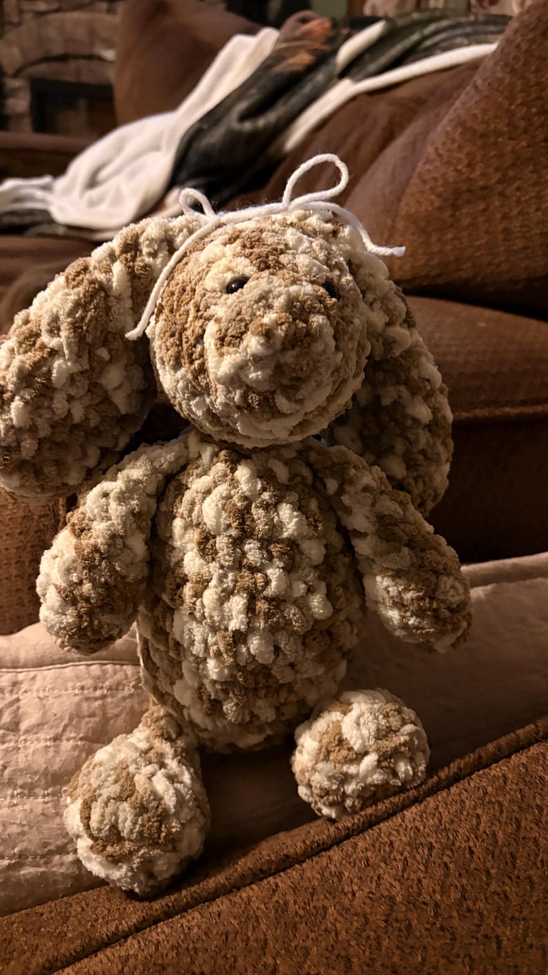 Op de afbeelding: Een handgemaakt, pluche konijnenspeelgoed in bruine en cr&egrave;mekleurige tinten. Het konijn heeft slappe oren, een rond lichaam en een lieve uitdrukking. Het speelgoed is gemaakt van zacht, getextureerd garen en heeft een wit touwtje om zijn hoofd.