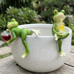May include: A white speckled ceramic planter with two green frog figurines. One frog holds a glass of red wine, the other a glass of yellow liquid. The frogs are perched on the planter's edge, creating a whimsical garden accent.