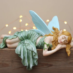 May include: A figurine of a sleeping fairy with light blue wings and long, wavy, golden hair. The fairy is wearing a green dress and is resting on a wooden surface. The fairy has pointed ears and is in a relaxed pose.