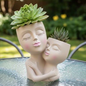May include: A beige ceramic planter shaped like two embracing faces, each with closed eyes and pink accents. The larger planter holds a green succulent, while the smaller one contains a spiky succulent. The planters are on a glass table.