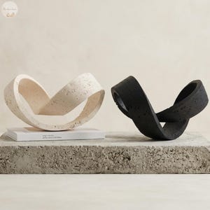 May include: Two abstract sculptures, one cream and one black, on a concrete block. The sculptures have a continuous loop design. A book with the text "weigh I had the perfect place to fall asleep" is visible.