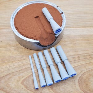 May include: A metal container filled with brown powder, with a carved-out section. Several light blue, cone-shaped tools are arranged around the container. The tools have a white body and a blue tip. The text "L 29MM" is visible on one tool.