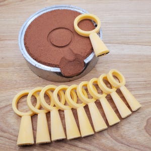 May include: Pale yellow ring-handled tools with a conical base, arranged on a wooden surface. A metal container filled with brown powder is in the background. The tools are likely for a specific craft or hobby.
