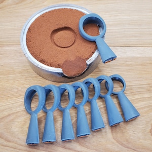 May include: A metal container filled with brown powder, next to several blue, handle-shaped tools. The tools have a circular handle and a conical base with a textured orange surface. The tools are arranged on a wooden surface.