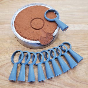 Op de afbeelding: Een verzameling blauwe ringvormige gereedschappen met conische handvatten, gerangschikt op een houten oppervlak. Een metalen bak bevat een bruin poeder, met een gereedschap erbovenop. De gereedschappen lijken bedoeld voor knutselen of het maken van sieraden.