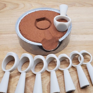 May include: A circular metal casting flask filled with reddish-brown sand, featuring a circular impression in the center. One white ring-shaped mold rests on the sand, while seven additional white ring-shaped molds are arranged in a row below.