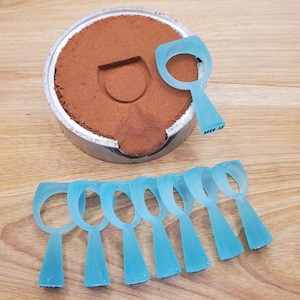 May include: A collection of light blue ring molds, with a size 12 marking, arranged on a wooden surface. A metal container filled with brown powder sits nearby, with a ring impression in the powder.