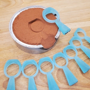 May include: A close-up shot of a metal container filled with brown powder, with a circular indentation. Several light blue, spoon-shaped objects are scattered around the container on a wooden surface. The objects have a flat, rectangular base.