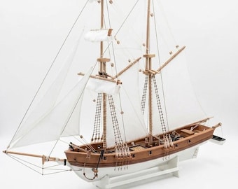 May include: A detailed wooden model of a sailing ship with two masts and white sails. The ship has a brown hull and deck, with intricate rigging and a white base. The model is a miniature replica of a classic sailboat.