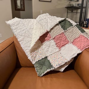 May include: A patchwork quilt in shades of white, pink, green, and floral patterns, draped over a brown leather armchair. The quilt's edges have a frayed, textured finish. The quilt is made of various fabrics and has a handmade appearance.