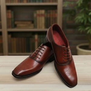 May include: A pair of brown leather dress shoes with a classic design. The shoes feature a lace-up closure, a square toe, and decorative perforations. The shoes are displayed on a light-colored wooden surface.