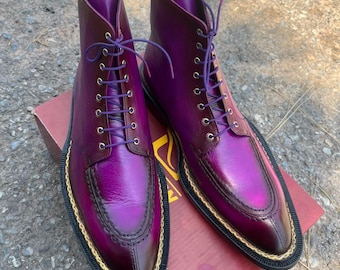 May include: A pair of purple leather boots with a high-top design. The boots have a lace-up closure with purple laces, a rounded toe, and a gold trim around the sole. The boots are displayed on a brown box.