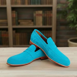 May include: A pair of vibrant turquoise suede loafers. These shoes feature a classic penny loafer design with a brown sole and trim. The background is blurred, showing a wooden surface and a bookshelf.