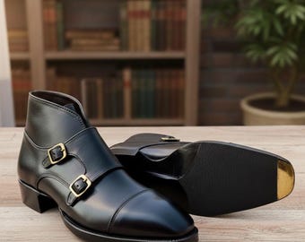 May include: A pair of black leather ankle boots with double monk straps and gold-colored buckles. The boots have a cap toe design and are displayed on a wooden surface. The sole of one boot is visible, showing a black rubber sole with a gold accent.
