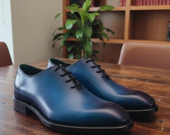 Puede incluir: Un par de zapatos de vestir de cuero azul con cordones negros, sobre una mesa de madera. Los zapatos presentan un efecto degradado, pasando de un azul más oscuro en la puntera a un tono más claro en la parte superior. Diseño Oxford clásico. Miden unos 30 cm de largo.