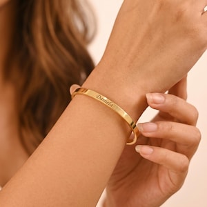 Könnte beinhalten: Ein goldfarbenes Armreif-Armband mit dem eingravierten Namen "Daniela". Das Armband wird an einem Handgelenk getragen und zeigt sein schlichtes, elegantes Design. Das Metall hat eine polierte Oberfläche.