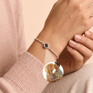 Könnte beinhalten: Ein silbernes Armband mit einem kleinen, runden, dunkel gefärbten Anhänger. Das Armband wird am Handgelenk getragen. Der Anhänger scheint ein Foto zu sein. Das Armband besteht aus kleinen, runden Perlen. Der Hintergrund ist ein zartes Rosa.