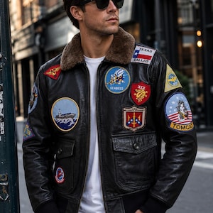 Op de afbeelding: Zwarte leren bomberjack met meerdere patches, waaronder "Texas Dallas" en "Tomcat". De jas heeft een bruine bontkraag en twee voorzakken. De persoon draagt een wit t-shirt en een donkere zonnebril.