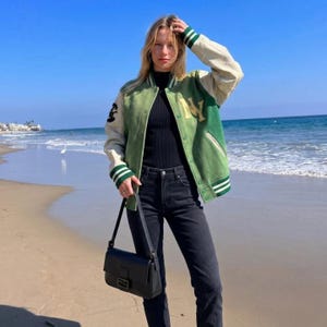 May include: A green and cream varsity jacket with the letter "NY" on the front. The jacket has green and white striped cuffs and trim. The person is wearing a black top and black jeans, and carrying a black shoulder bag.