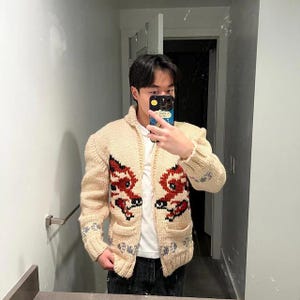 May include: Cream-coloured, hand-knitted cardigan with a red and black dragon design on the front. The sweater has a zip-up closure, pockets, and a high collar. The sleeves and bottom hem feature a grey and blue pattern.