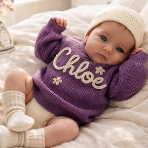 May include: A baby wearing a purple sweater with the name "Chloe" in white script, accented with a small daisy. The baby also wears a cream-colored beanie and socks. The baby is lying on a bed with a white pillow.