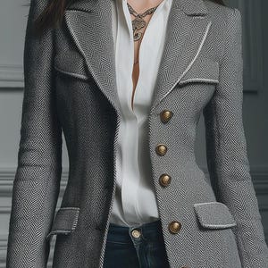 Puede incluir: Una blazer gris con estampado de espiga con botones dorados y ribete blanco. La blazer se lleva sobre una camisa blanca con cuello y vaqueros azules. Se ve un collar dorado. La blazer tiene dos bolsillos delanteros.