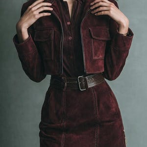 May include: A burgundy suede outfit featuring a cropped jacket with pockets, a button-down shirt, and a matching skirt. The outfit is accessorized with a brown leather belt with a silver buckle. The ensemble is a stylish, coordinated set.